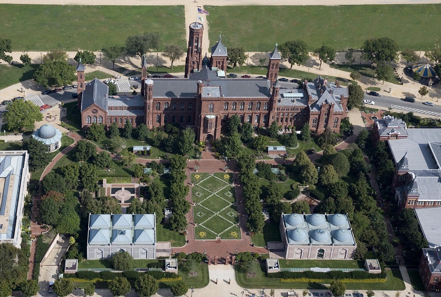an aerial view shows the red brick Smithsonian castle with a formal garden behind it and several other structures around it