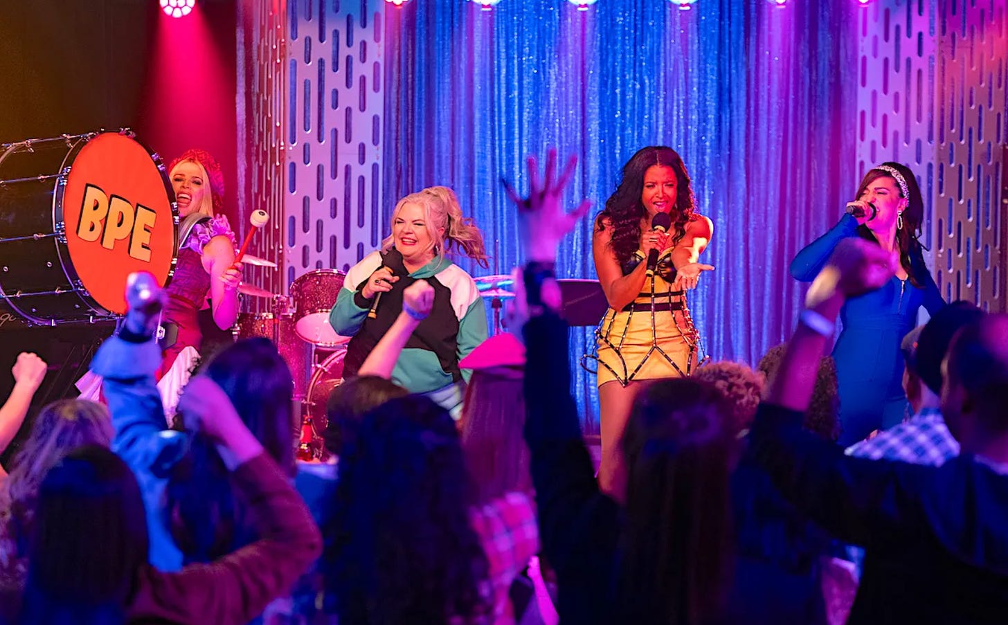 Busy Philipps, Paula Pell, Renee Elise Goldsberry and Sara Bareilles in Girls5eva, performing a pop concernt on stage in brightly colored attire