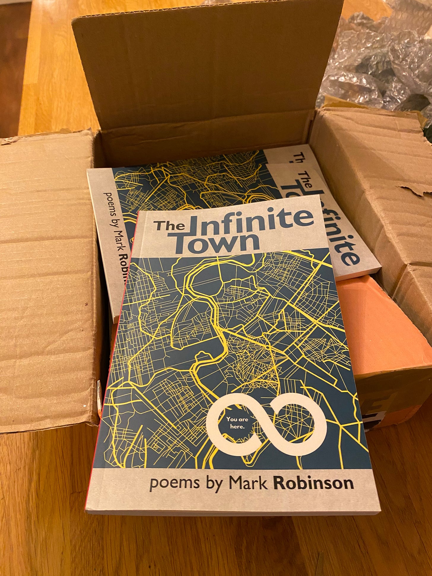 Paperback copies of The Infinite Town poems by Mark Robinson in a just opened package. Green and yellow map with infinity sign - in one half of which it reads: You are here.