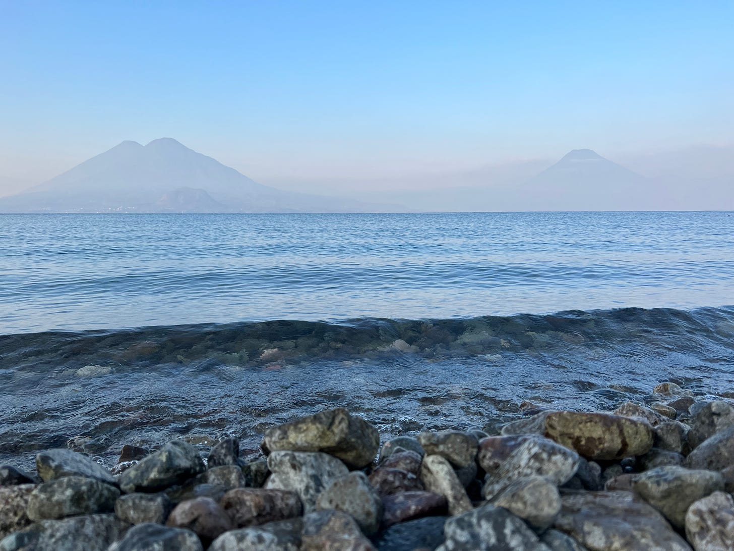 a lake in the morning with two volcanos shrouded by haze in the background set against a blue sky