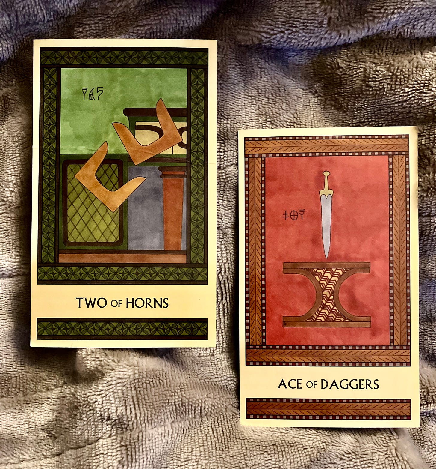 Two Minoan Tarot cards side by side on a grey velveteen background. The Two of Horns is in shades of green and brown. It shows two pairs of Minoan sacred horns tumbling off a shrine. The Ace of Daggers is in shades of deep red and tan. It shows a large gold-hilted dagger hovering, point downward, over an incurved altar.