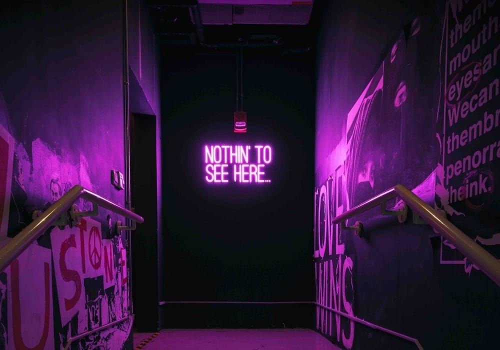 Neon purple sign at the end of bar hallways with graffiti saying "Nothin' to see here."