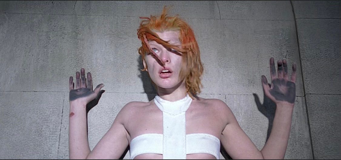 A young woman with matted orange hair, dressed in an outfit of bandages that strategically cover her intimate bits raises her grime-coated palms in acquiescence against a stark concrete wall. She has no file because she is Leeloo, the Fifth Element.