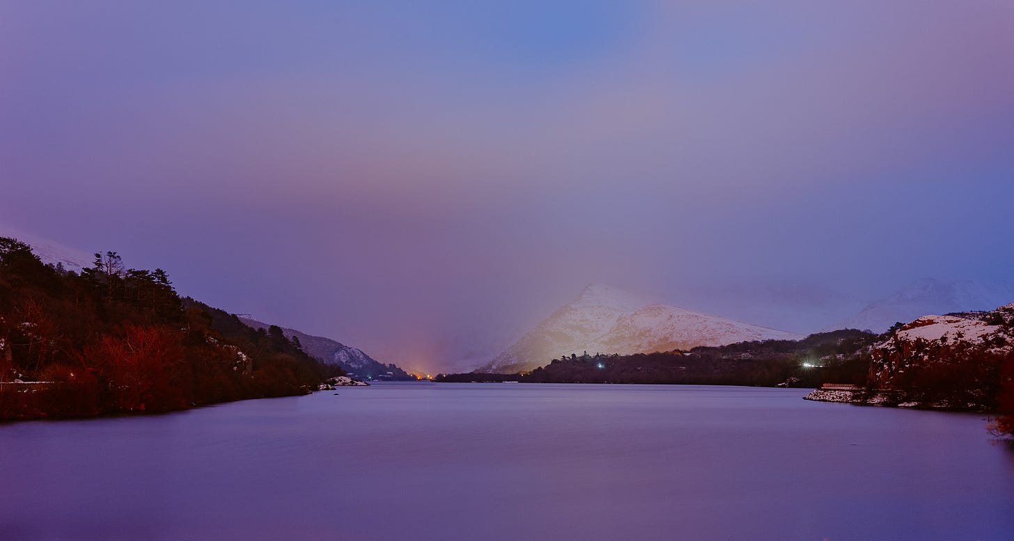 At dusk the sky is purple and reflected in an ice-covered lake. In the distance there are mountains covered in snow.
