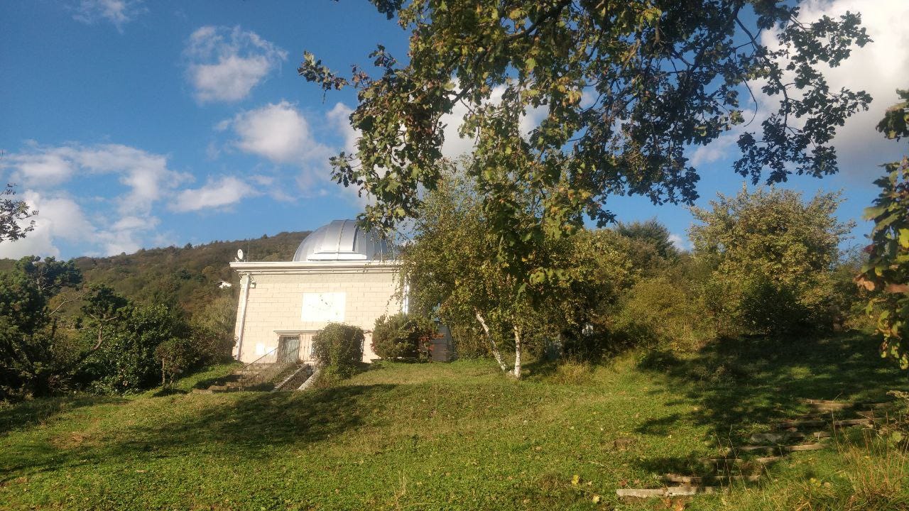 Vista di un prato in montagna con un albero in primo piano sulla destra, cielo azzurro con nuvole bianche e nel centro un osservatorio astronomico con una cupola grigia. La foto è scattata di giorno, con luce del sole che si riflette sulla parete frontale bianca dell'osservatorio.