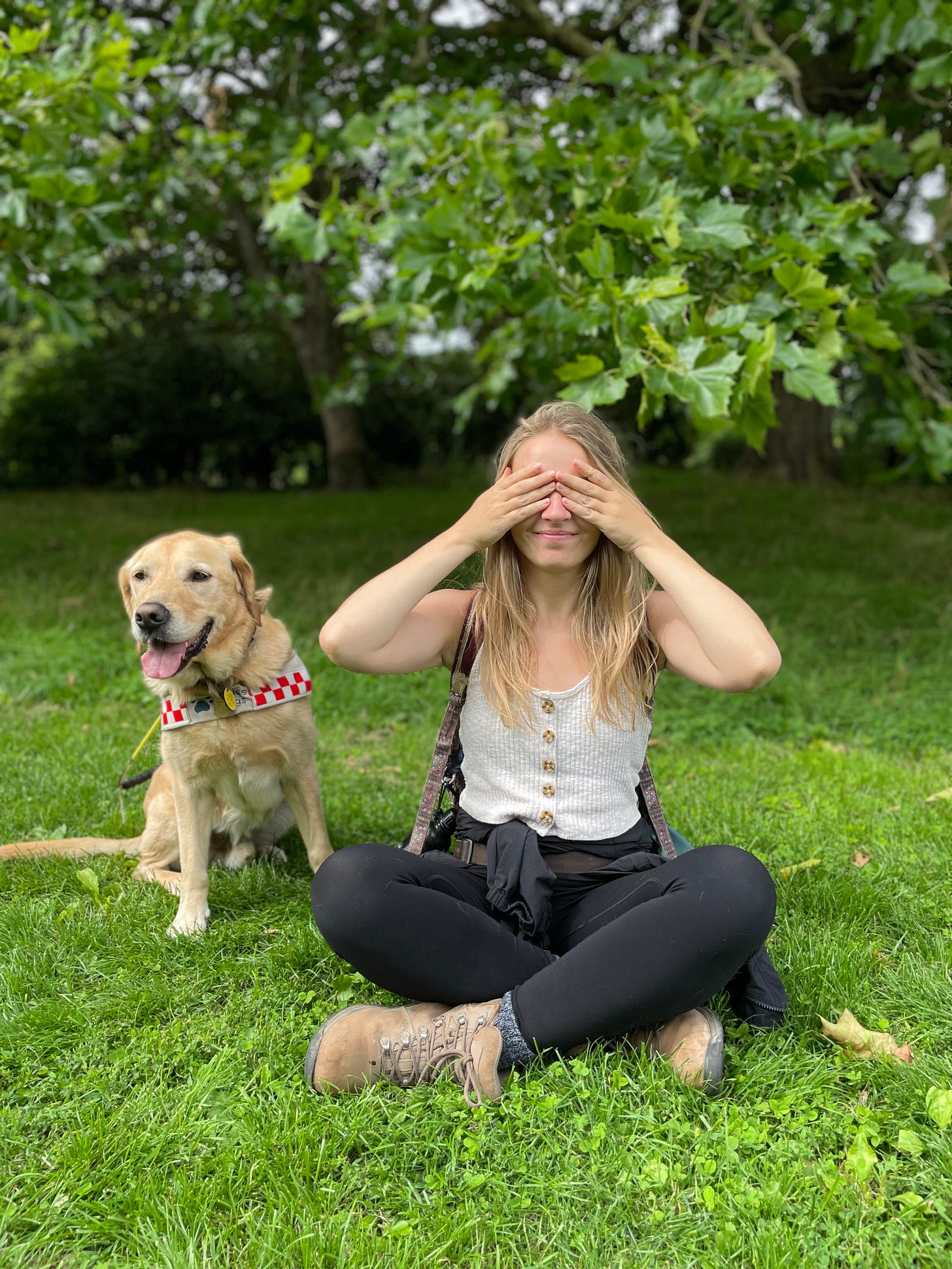 A photo of me and my guide dog sitting on a grass with lots of leaves visible in the background, making the photo look very greenery. My legs are crossed and I am covering my eyes with my hands.