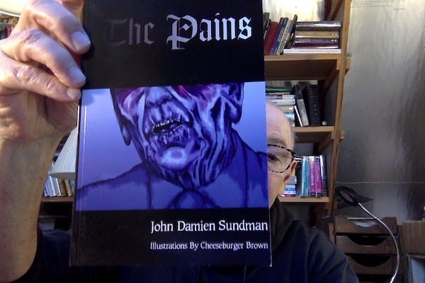 Picture of some old bald dude holding a copy of my novella The Pains ("By John Damien Sundman, Illustrations by Cheeseburger Brown") which features the title in gothic text, in which the word 'The' in 'The Pains' is fading to black, barely visible. under the title there is a nightmarish illustration, in dark blue, of the bottom 2/3s of Ronald Reagan's face.