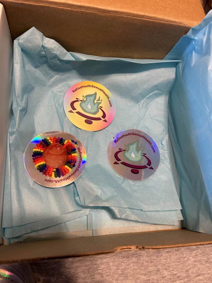 A box lined in light blue tissue paper, three round stickers: one with a beaded rainbow condom on a holographic background, one with the NYSHN logo of a turtle carrying a fire on holo background, and one of our logo with a soft pastel rainbow background