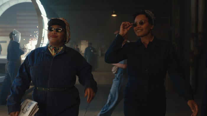 Two Black women are confidently sauntering side-by-side through an active factory. They are smiling and wearing dark blue work coveralls, stylish head scarves, and dark sunglasses. The woman on the left is carrying papers and the woman on the right is adjusting her sunglasses.