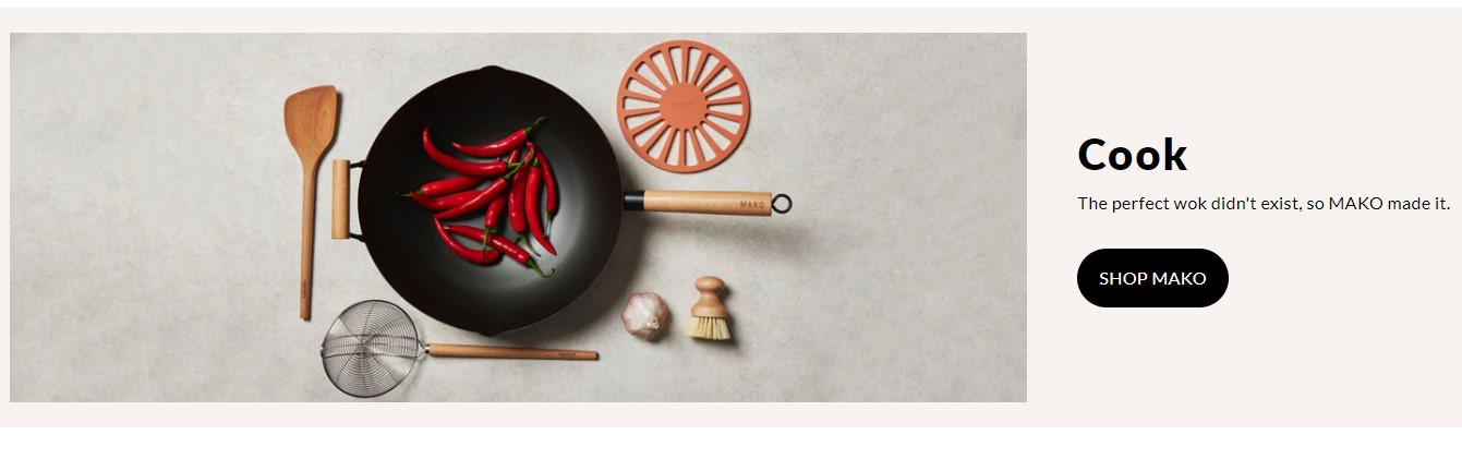 An image from the Mako website. An overhead shot of a wok containing whole red chiles. Beside the wok are a trivet, a spider strainer, a spatula, a head of garlic, and a scrub brush. Copy beside the photo reads "Cook. The perfect wok didn't exist, so Mako made it."