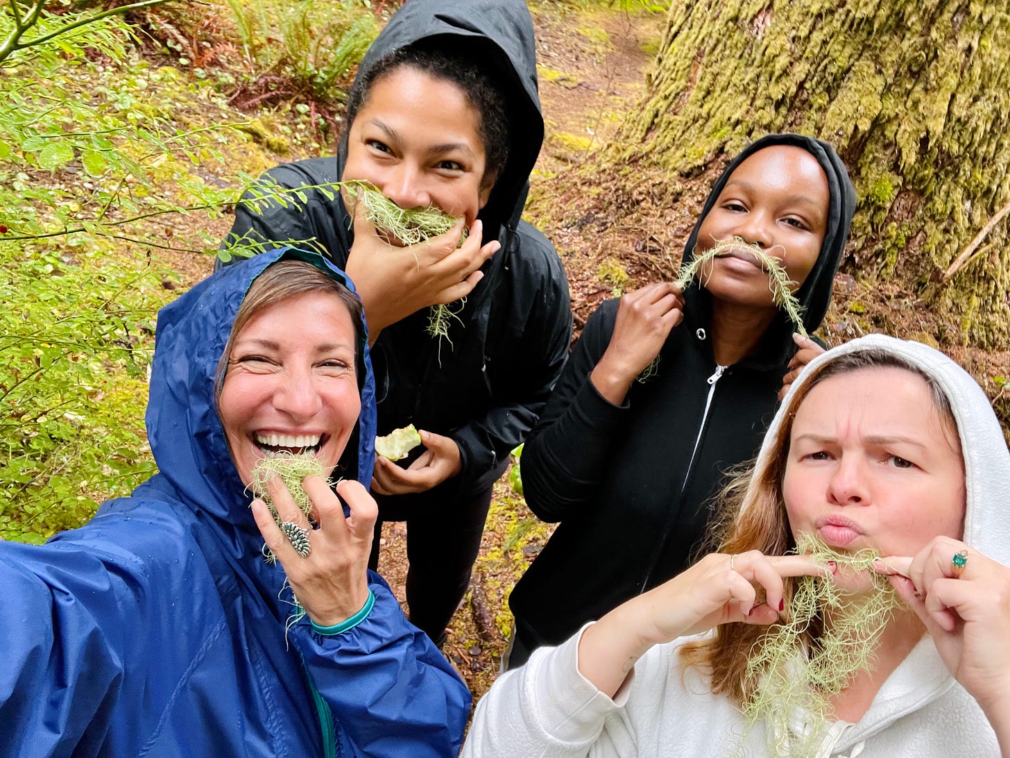 Nicole, Amber, Jackie, and Nafissa stand on a trail in the woods on a rainy day. Nicole is taking a selfie of the group. They each hold parts of a plant up to their faces as if the green hair-like plant is facial hair. 