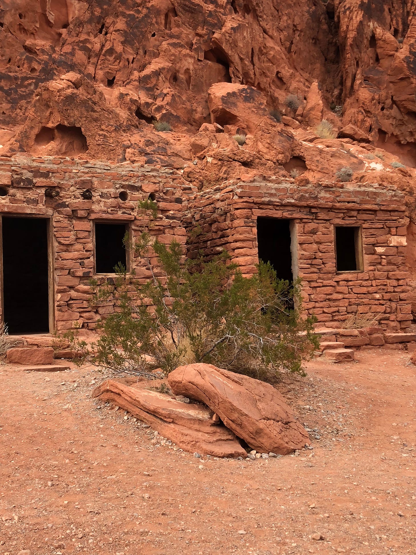 Two adobe brick buildings at the base of a red rock mountain. Each building has an open door and window. in front of the buildings grows a small tree with green leaves behind 2 red rocks, one atop the other.