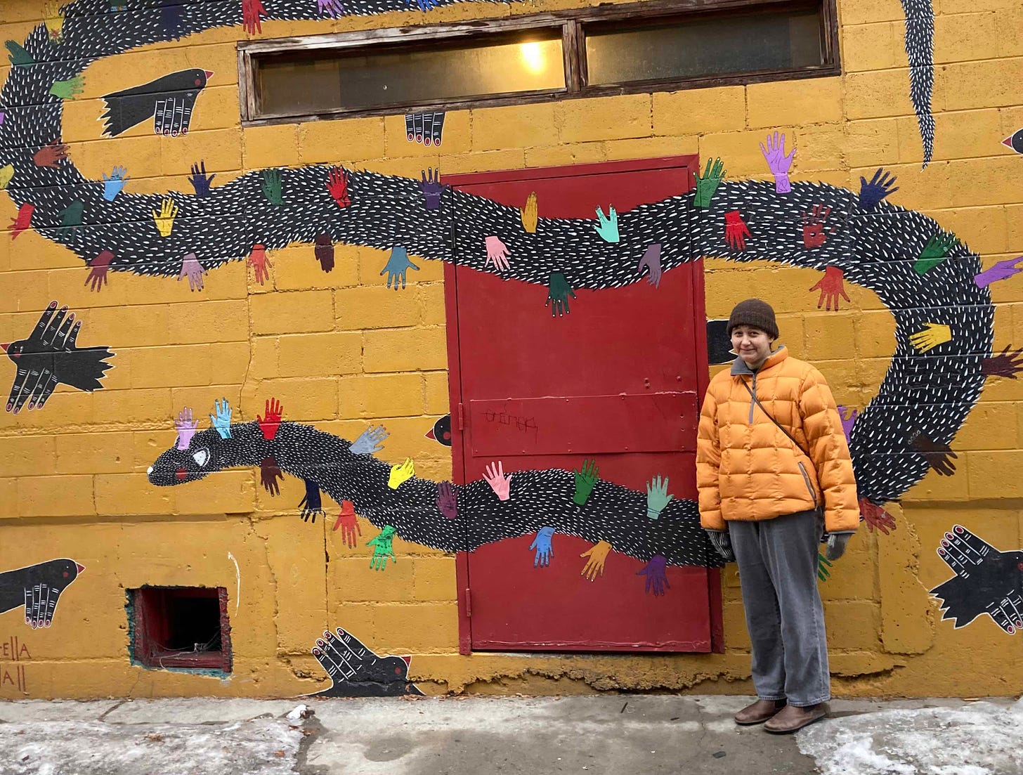 woman in puffy jacket and hat in front of bright mural