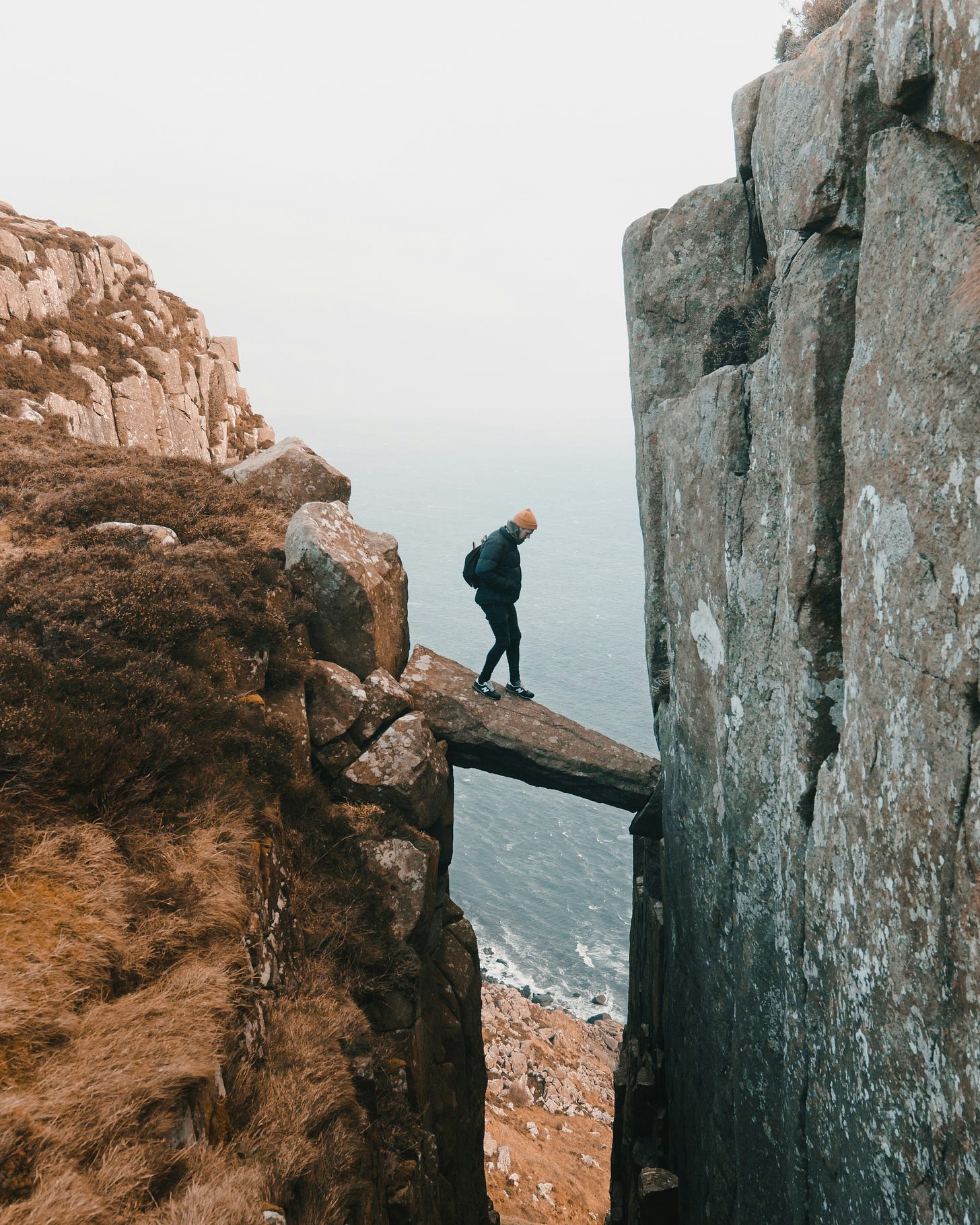 The image shows a person standing on a rocky cliff overlooking a body of water, likely an ocean or sea. The cliff appears to be quite high and steep, with jagged rocks and a misty, overcast atmosphere. The person is dressed in dark clothing, suggesting they may be exploring or hiking in this remote, rugged natural landscape.