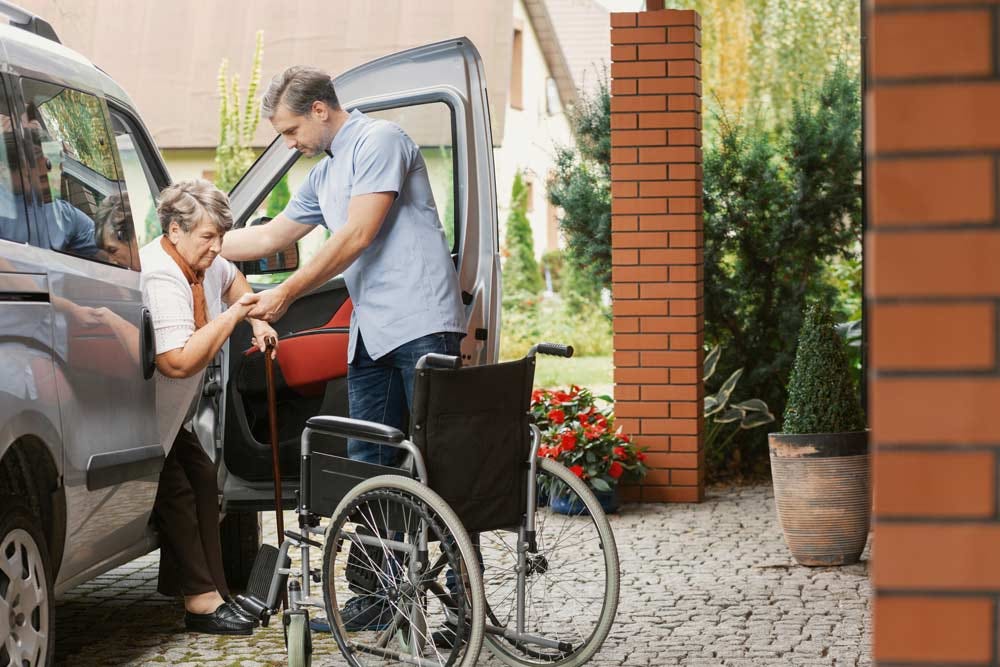 Son helping elder parent out of a car & into a wheelchair.