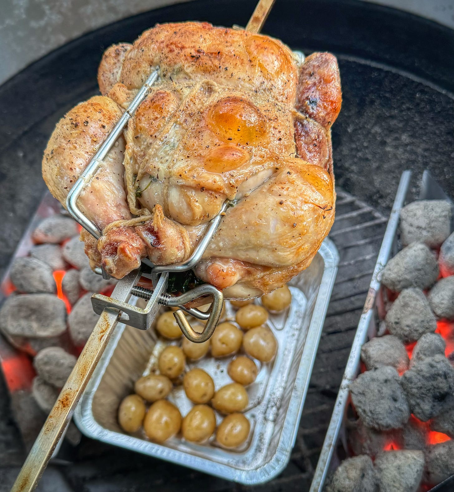 A chicken on a rotisserie on a charcoal grill. The skin of the chicken is brown and there are puffed-up bubbles in some places. There is also a drip pan full of roasting potatoes below the chicken. 