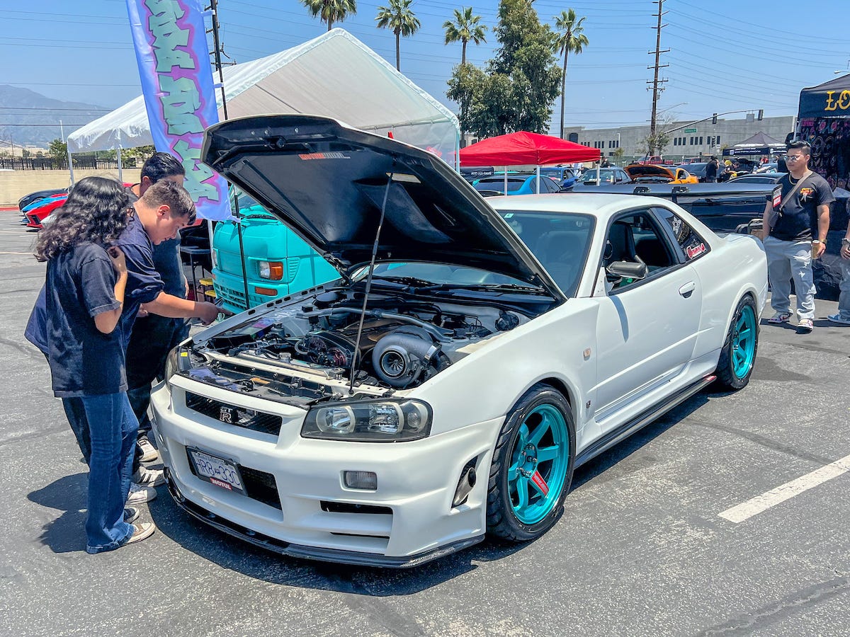 A while Nissan R34 Skyline GT-R with teal wheels and extensive aftermarket modifications to the engine bay and bodywork seen at Fuelfest Los Angeles in June 2023 at Irwindale Speedway.
