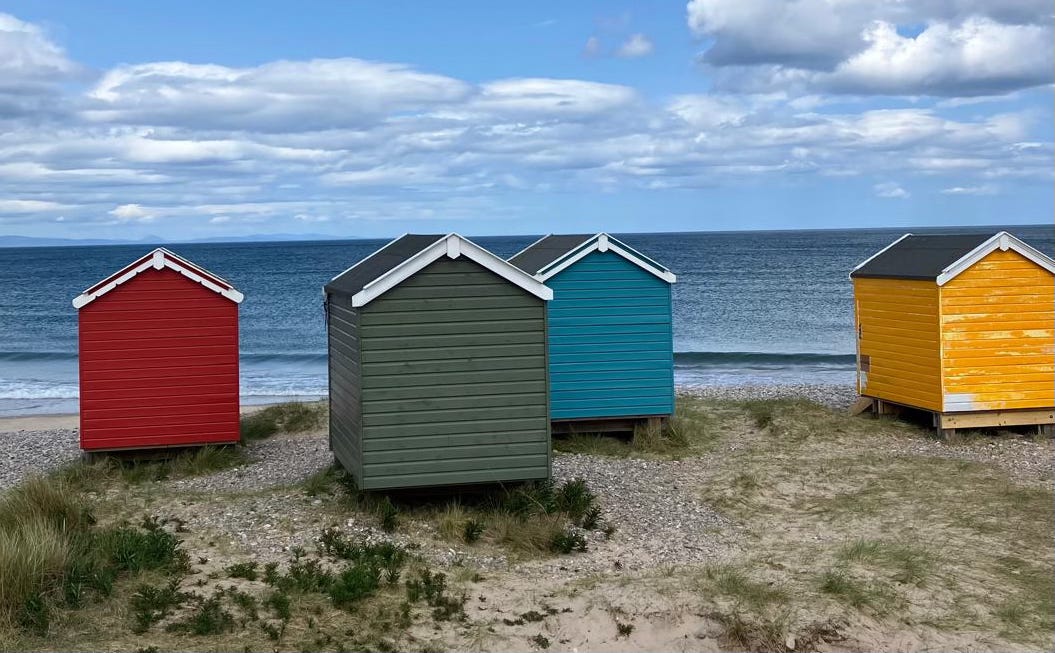 Four brightly colored wooden beach huts—red, green, turquoise, and yellow—stand on grassy dunes overlooking the calm sea near Findhorn on the Moray Firth in Scotland. The sky is partly cloudy, and gentle waves lap at the shoreline in the background.