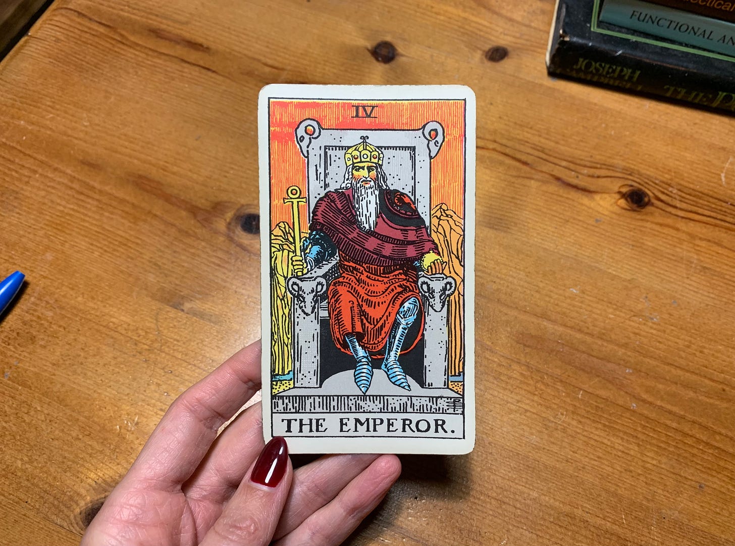 A hand is holding a Tarot card, The Emperor by Pamela Colman Smith. In the image, a person is wearing all red with armor underneath. He is wearing a crown and sitting on a throne with ram's heads on each corner. There are mountains in the background and a thin river flowing. Behind the card is a wooden desk with books on it and some pens.