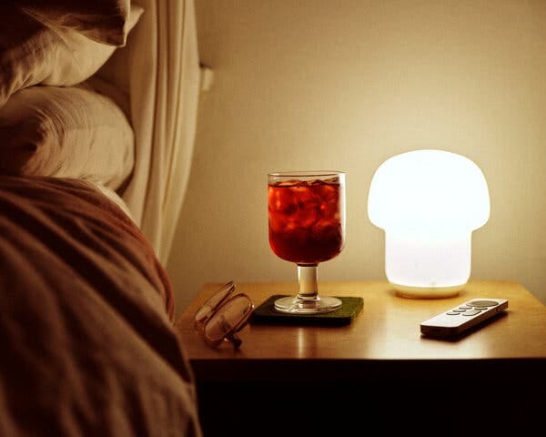 Image of bedside table with a clear glass with red colored mocktail, a bedside lamp, eyeglasses and remote control. 