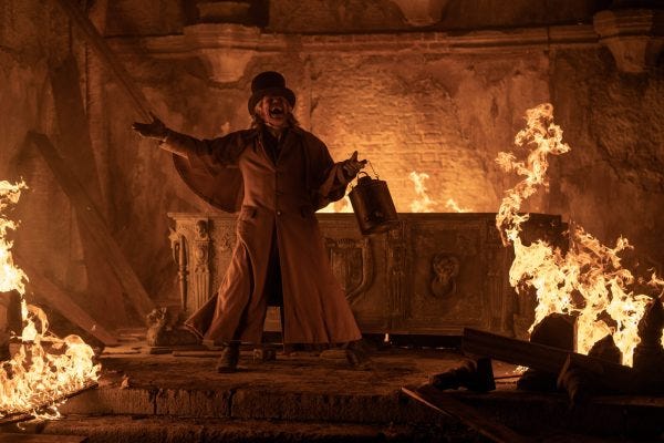 Willem Dafoe as Albin Eberhart von Franz gestures widely, holding a lantern and surrounded by flames