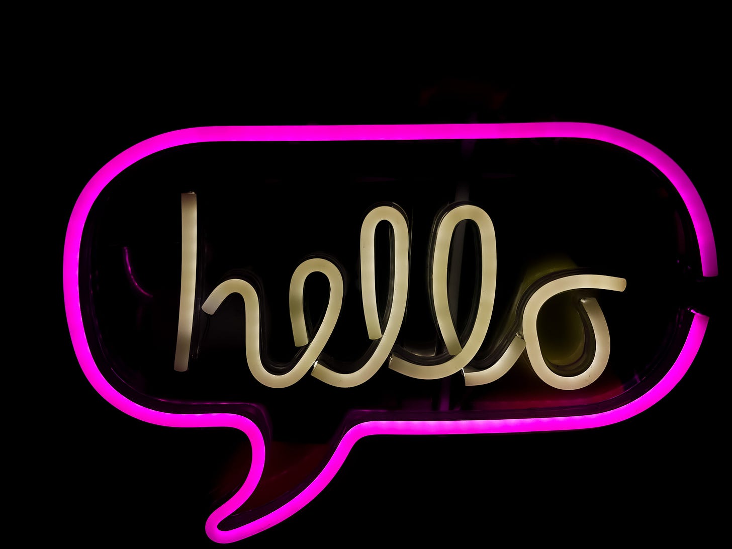 Neon pink speech bubble with the word hello written in brass cursive over a black background.