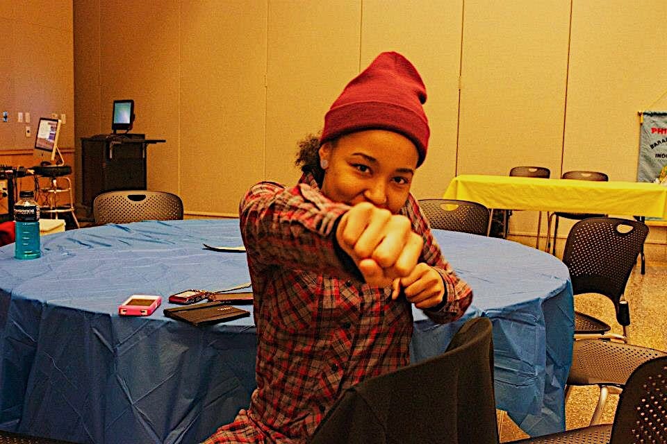 A black girl faking a punch at the camera with a burgundy beanie and plaid gray, black, and red shirt sitting in a black chair in a gymnasium set up for an event.