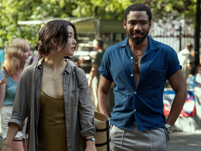 maya erskine and donald glover in mr. and mrs. smith, walking around a farmers' market. erskine is in a green slip dress and black denim overshirt, and glover is wearing fitted grey slacks, and a blue shirt unbuttoned slightly to show his pecs