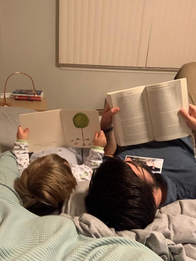 A toddler and a man read books together on a bed 