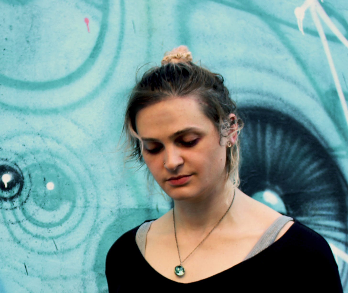 A picture of Jocelyn Burnham, a femme presenting person in her mid 30s, pictured against a creatively painted wall and wearing a black top, planet necklace and a nose stud with hair tied back in a messy bun.