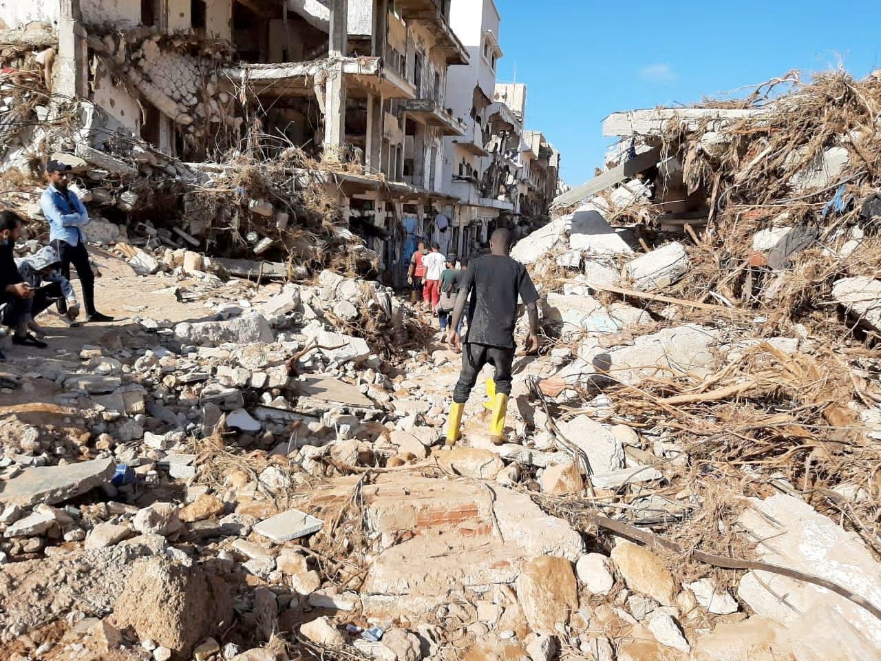 A boy in yellow boots navigates rubble on a Derma street.