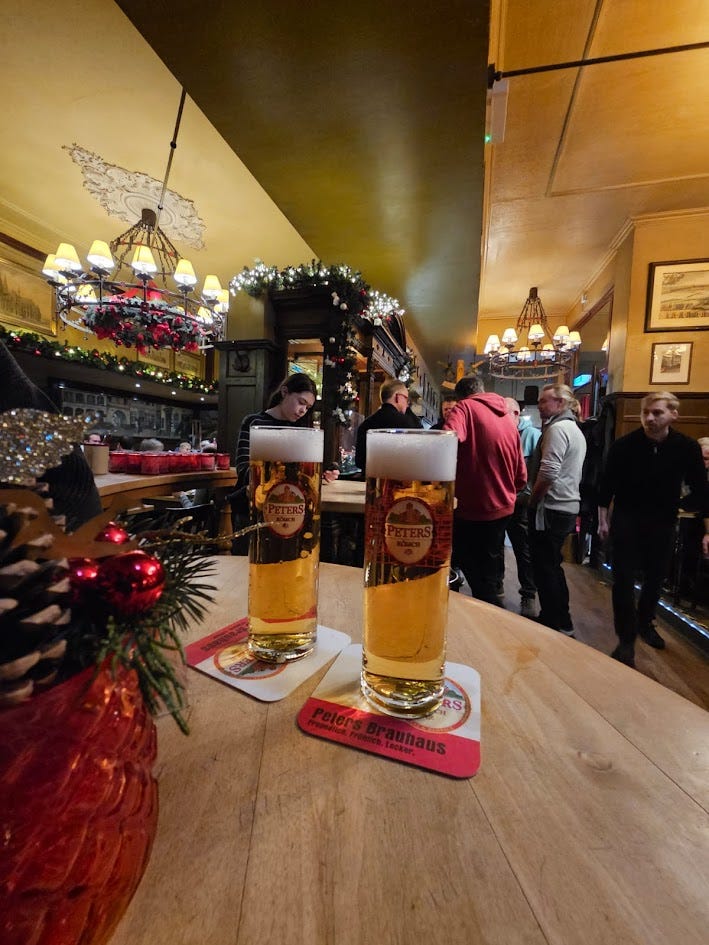 Two glasses of kolsch with the Peters logo printed on them in white and red. The setting is very festive, with shiny red baubles and pine cones and spruce decorating the tables and chandeliers.