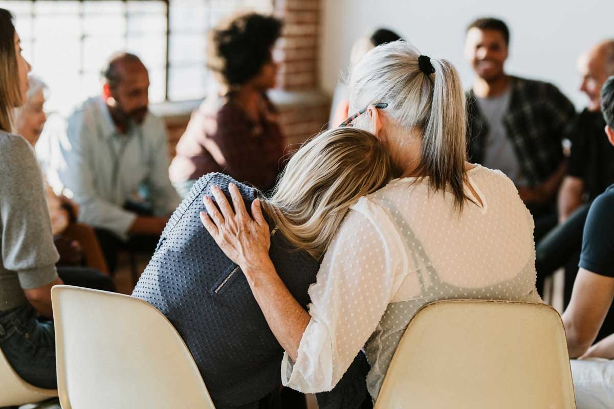 Young individual leans on an older woman’s shoulder for support during a group discussion.