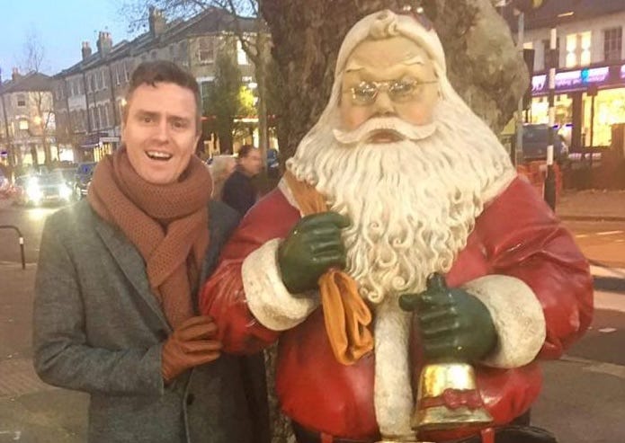 Me in 2016 standing next to a life-size figure of Santa who is ringing a bell. I am wearing a grey coat, tan gloves and an orange scarf which is quite large now I come to think of it