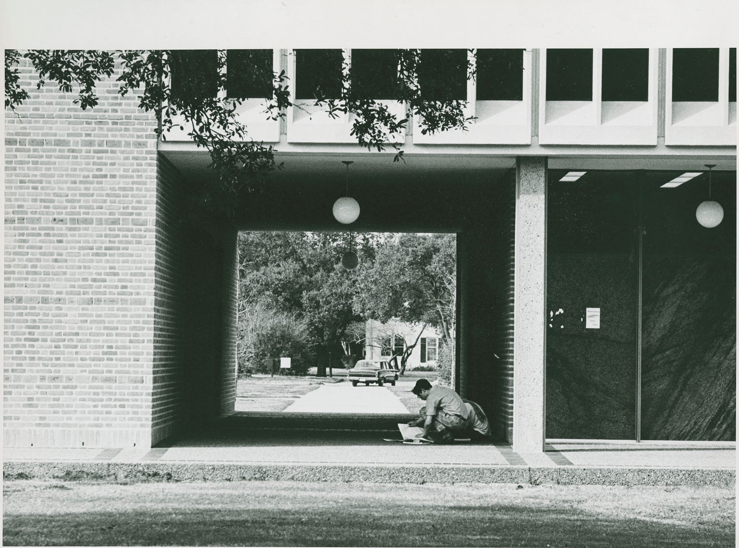 Black-and-white picture of the breezeway. The corridor is rectangular. There is a white 1960's-style globe light hanging from the ceiling. We can see a Ford Falcon through the breezeway, and a student is seated, studying