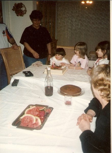 A photo from the 90s of a little girl in front of her birthday cake. Other family members are around the table, which has a white table cloth. There is another cake, a bottle of wine, and a tray of watermelon slices, too.