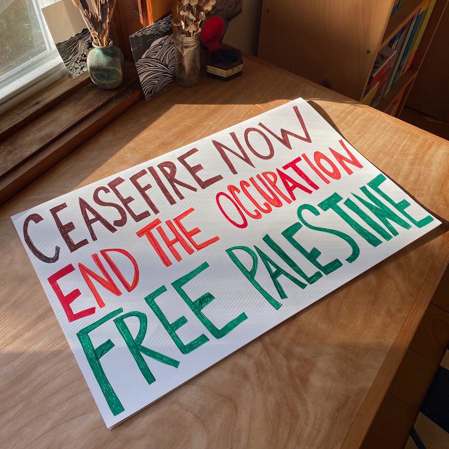 A white cardboard sign that says ‘CEASEFIRE NOW. END THE OCCUPATION. FREE PALESTINE.’ lies flat on a sunny wooden desk.