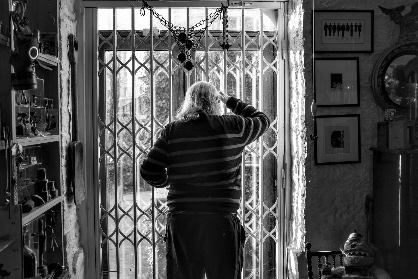 On a visit to see Wilf Lunn I took this photo of him looking out through a locked door with a grill on it. The photo has his back to the camera almost in silhouette with his hand up to shed his eyes. The shelves and walls on either side are cluttered with knick knacks.
