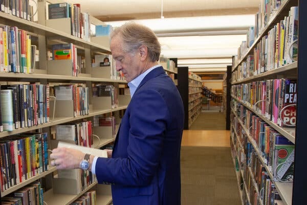 A middle-aged man in a blue suit peruses a book in the middle of a library, with shelves of books on either side of him.