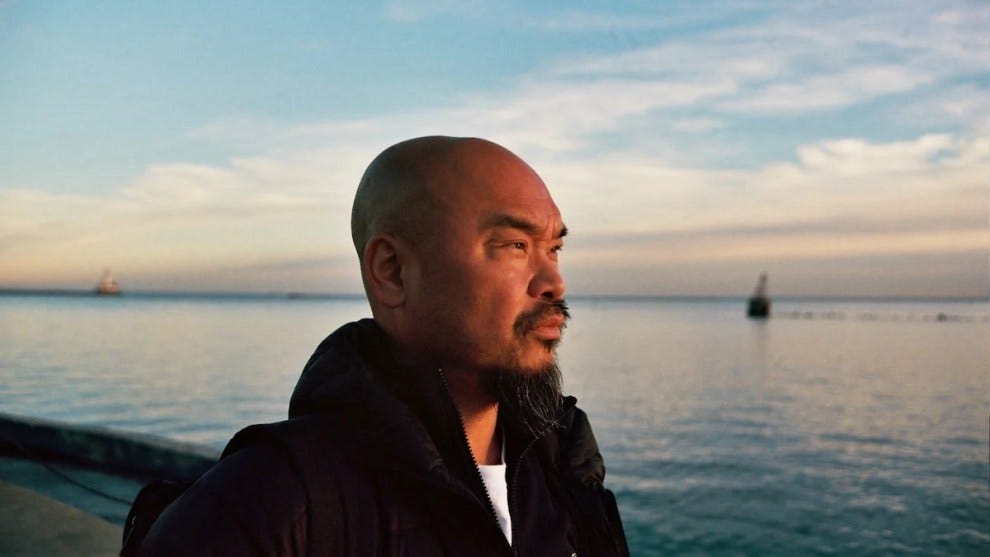A bald man with medium-toned skin with a body of water in the background, looking out to the distance on the right.