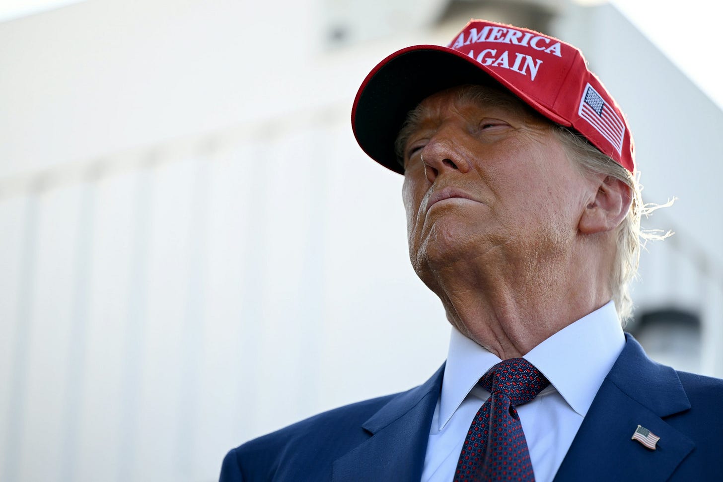 Former President Donald Trump wearing a red 'Make America Great Again' hat, looking upwards with a serious expression, in front of a blurred background.