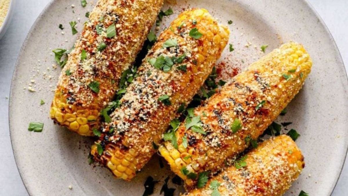 Grilled corn on the cob generously seasoned with spices, herbs, and cheese, presented on a round, light gray plate.
