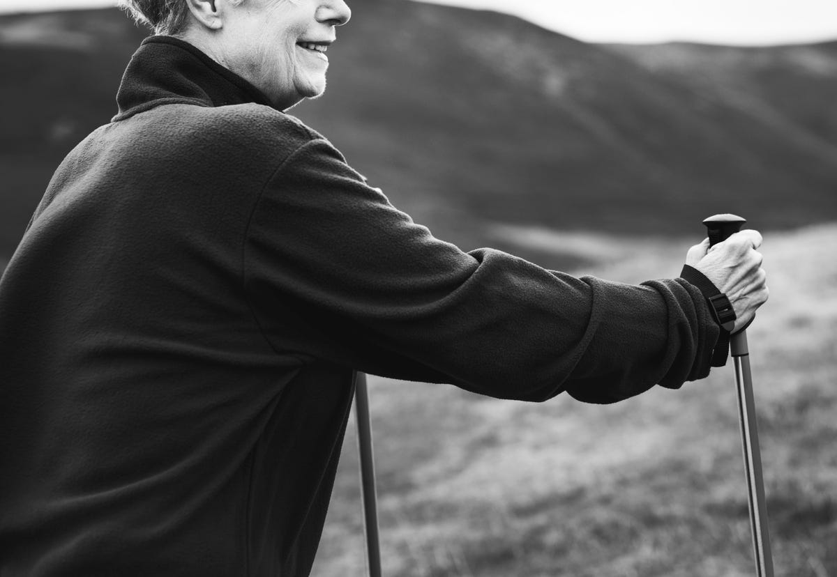 Black and white photo of a middle aged woman smiling, walking with walking poles on a hill.