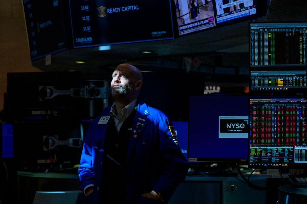 A slice of light from above illuminates a bearded man's face as he gazes upward, computer monitors full of red and green stock data buzzing behind him.