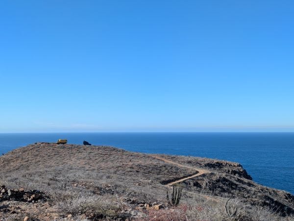 two tiny looking trucks out at the end of a desert landscape with the blue ocean and a cloudless blue sky in the background