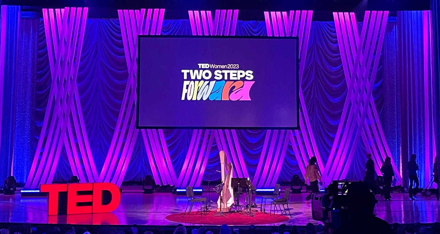 A photo of the TEDWomen stage with the big TED letters on the left, a stage set up with band instruments in the center, and a screen above that reads 'TEDWomen2023: Two Steps Forward'