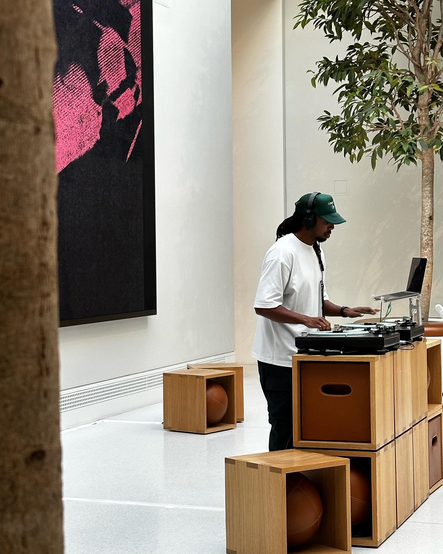 A DJ performs in the Forum at Apple Carnegie Library. Forum cubes are stacked to make a table, and DJ equipment sits on top. In the foreground and background, there are trees.