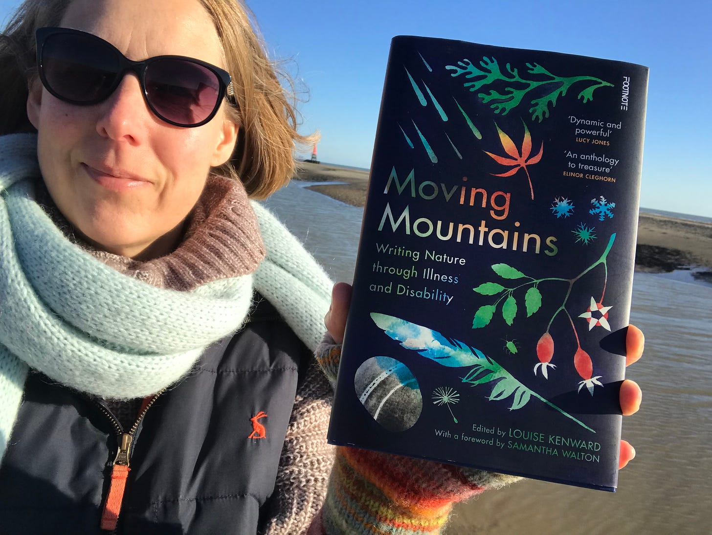 photo of me, a white woman in sunglasses and a pale blue wooly scarf holding up a copy of Moving Mountains on the beach, water and sandy stretch behind me below a bright blue sky.