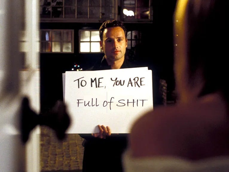 An image from the movie Love, Actually. A man whose character name I can't be bothered to look up, but you know the one, he's holding the cards with the hand written messages. The message on the card he's holding says "TO ME YOU ARE FULL OF SHIT
