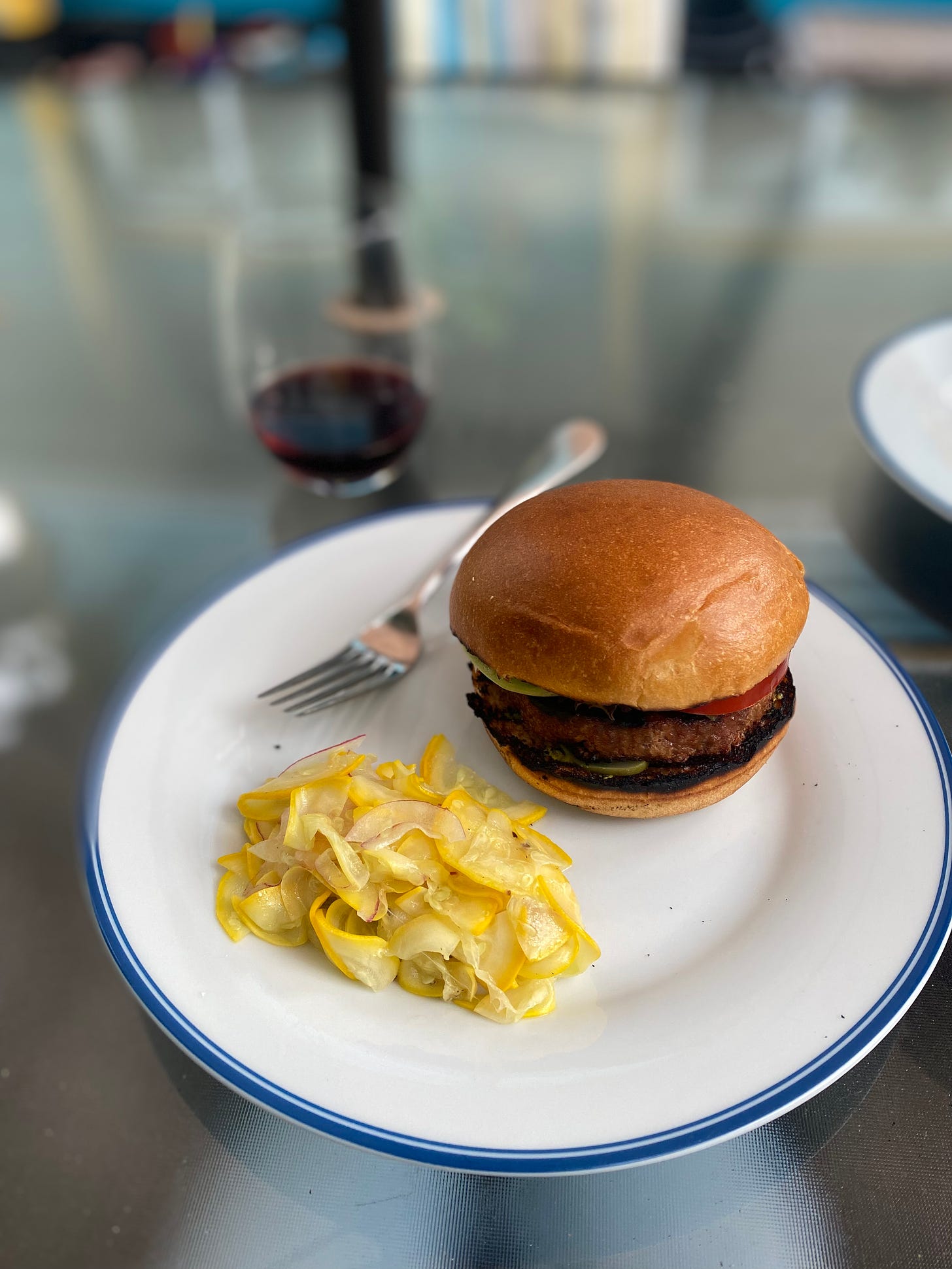 A white plate with a blue rim, holding a Beyond burger on a grilled bun and a shaved yellow zucchini salad. A fork rests on the plate at the back, and blurred in the background is a stemless glass of red wine.